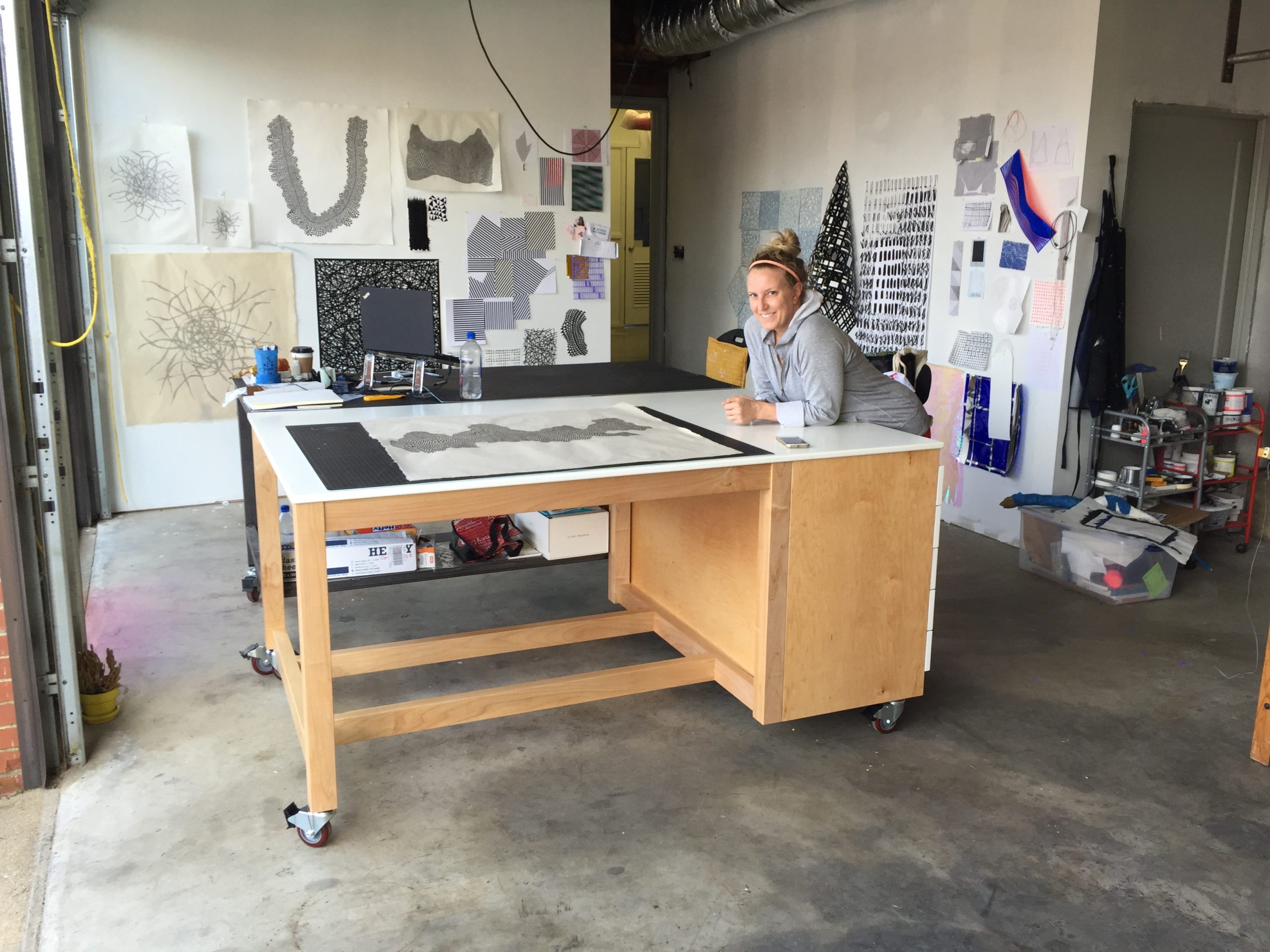 Female artist in an art studio interior with a workbench, art supplies, and various artworks displayed on walls.