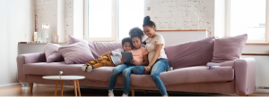 A woman and two children are seated on a couch, with the child in the center holding a tablet and all three looking at the screen together.