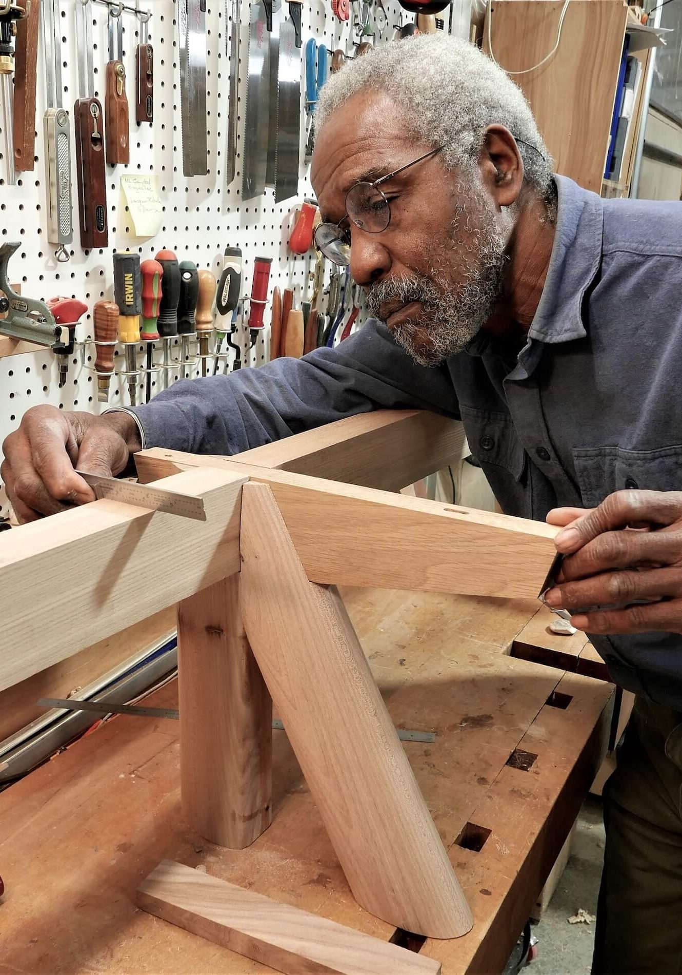 Older male artist measuring wood at a bench in a workshop full of tools hanging on a peg board
